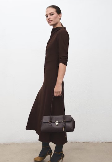 Žena u smeđoj dugoj haljini drži crnu torbu i stoji