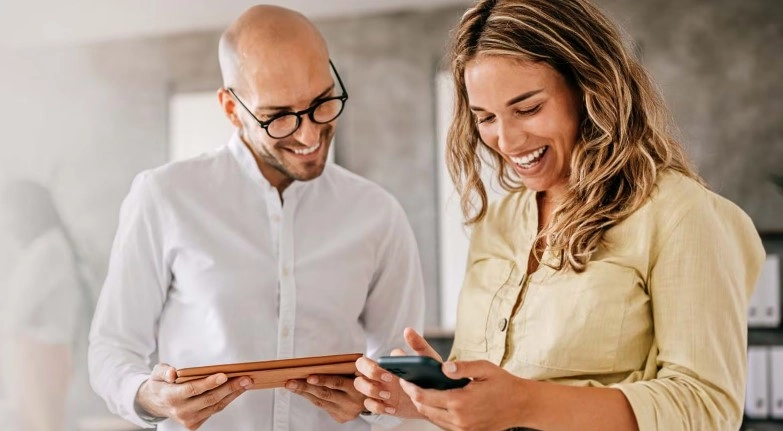 Dvoje ljudi gledaju mobitel i tablet i smiju se